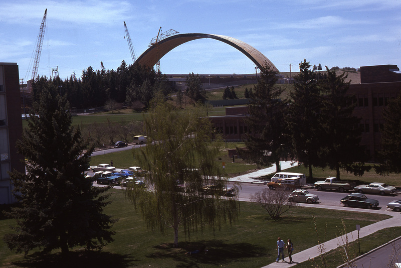 Color photograph of a grassy area surrounded by brick buildings. In the distance is a large arched roof under construction.