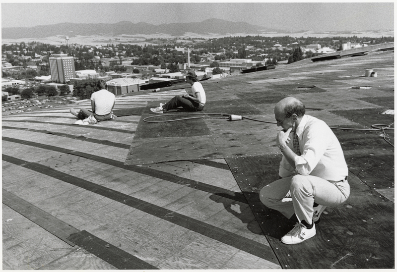 Black and white photograph of a man kneeling on the arched roof of a building. Two other people and some tools also sit on the roof. In the background are buildings and hills.