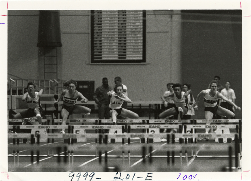 Black and white photograph of five runners jumping over hurdles that read "IDAHO". In the background are more people standing.