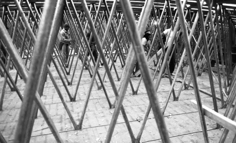 Black and white photograph of people walking among crossed vertical metal bars.