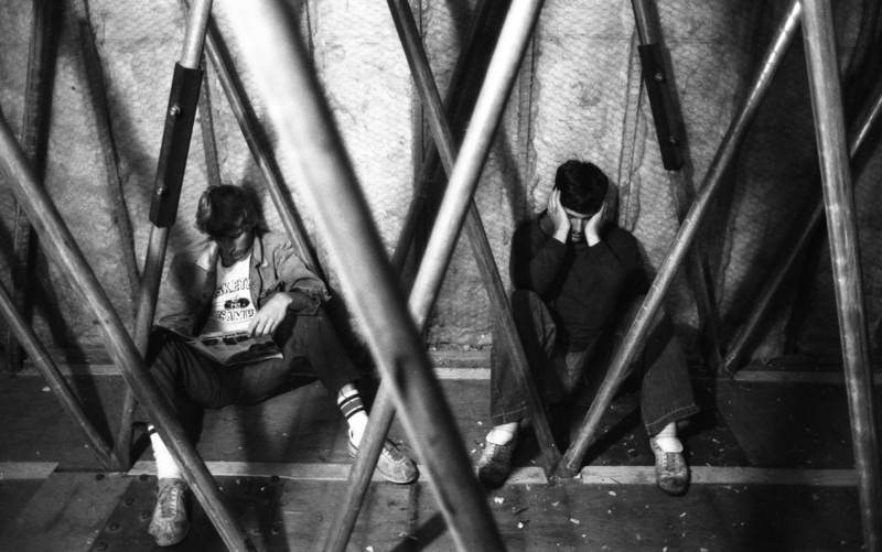 Black and white photograph of two people sitting against a wall among crossed vertical metal bars.