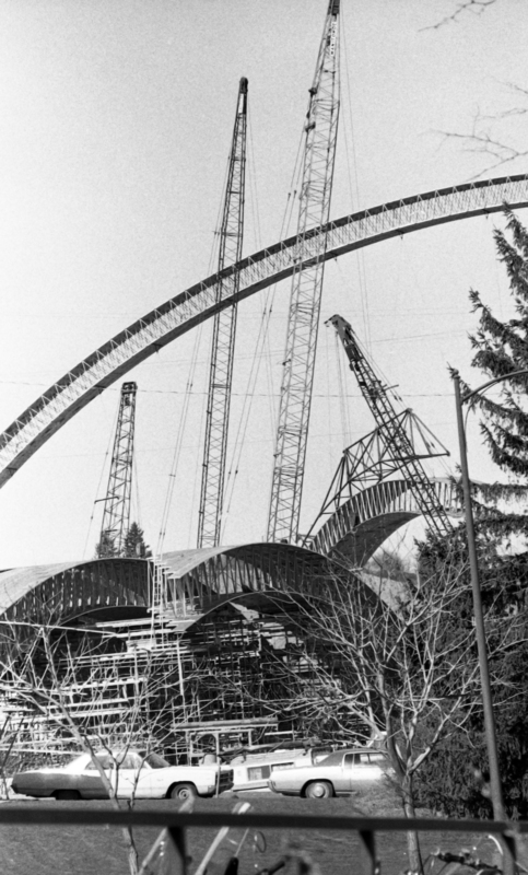Black and white photograph of a construction site. One roof arch rises above the ground, surrounded by construction cranes, and more wooden roof arches sit on the ground below.