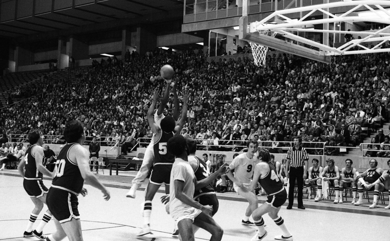 Black and white photograph of a basketball game in progress. In the background are stands filled with people.