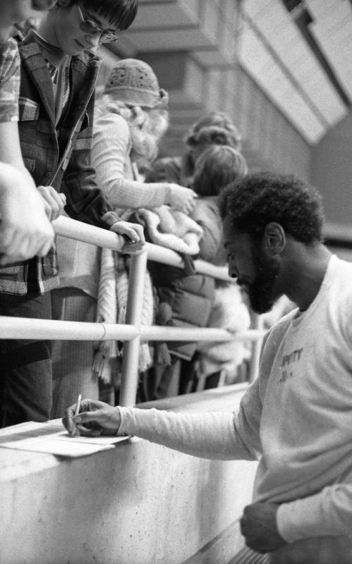 Black and white photograph of a man signing a piece of paper. People standing in the stands above him look down toward him.