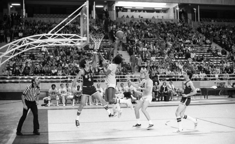 Black and white photograph of a basketball game in progress. People sit in the stands in the background.