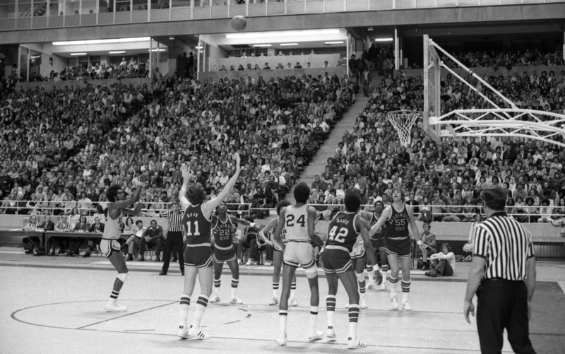 Black and white photograph of a basketball game in progress. People sit in the stands in the background.