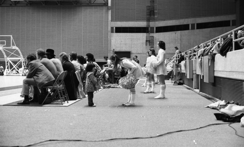 Black and white photograph of a young woman interacting with a child on the sidelines of a basketball court. Other people stand or sit in the background.