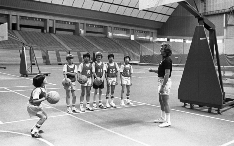 Black and white photograph of a man and a group of children on a basketball court in an enclosed stadium. One boy is about to throw a basketball toward a basketball hoop.