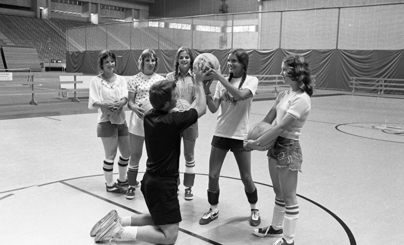 Black and white photograph of a man and a group of young women on a basketball court in an enclosed stadium. The man appears to be teaching of the young women how to hold a basketball properly.
