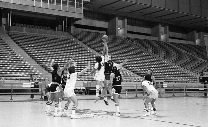 Black and white photograph of basketball players in two different uniforms, jumping for the basketball on a court in an enclosed stadium.
