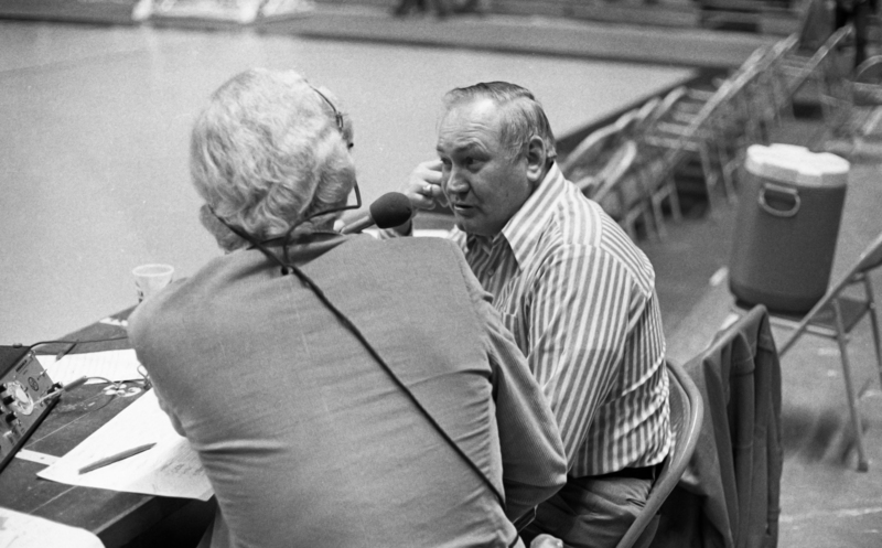 Black and white photograph of two men talking to each other, one holding a microphone to the other. In the background are folding chairs and a cooler.