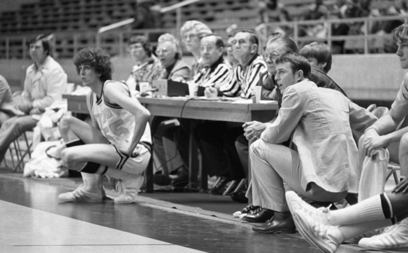 Black and white photograph of several people on the sidelines of a basketball court, two kneeling and the others sitting behind a table. In the background are stands.