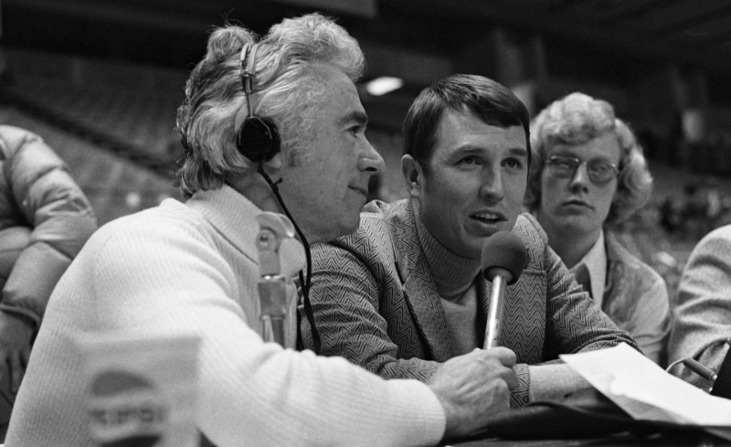 Black and white photograph of three men. One man, wearing headphones, holds a microphone out to one of the others.