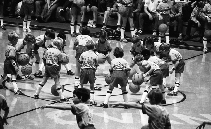 Black and white photograph of a bunch of children standing in a circle on a basketball court, dribbling basketballs.