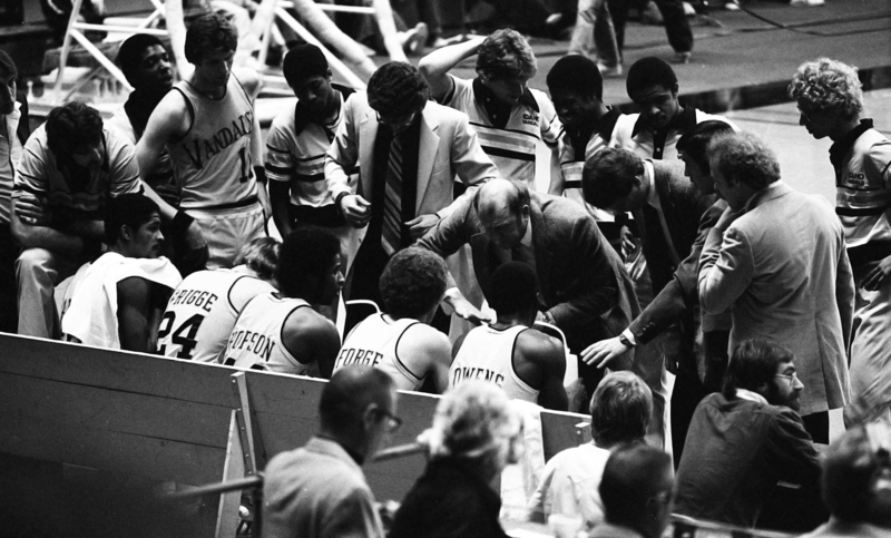 Black and white photograph of a man in a suit talking to a group of men in basketball uniforms on the side of a basketball court.