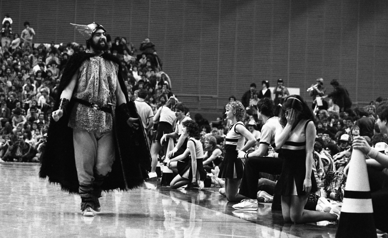 Black and white photograph of a man in a costume walking by cheerleaders kneeling by the side of a basketball court. In the background are people sitting in stands.