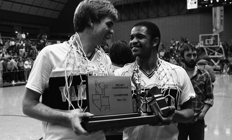 Black and white photograph of two men, basketball hoop nets draped around their necks, holding a plaque reading "BIG SKY CONFERENCE CHAMPIONS 1981-82". In the background are people sitting and standing on stands.