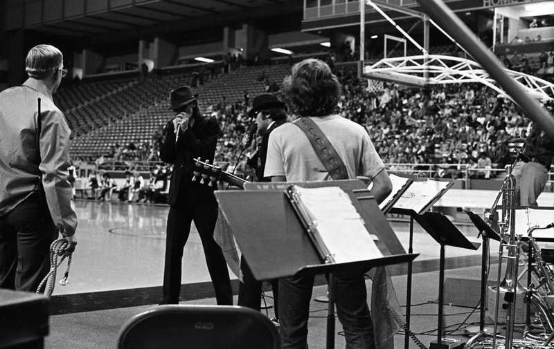Black and white photograph of a group of musicians playing at the end of a basketball court, two of them dressed in dark suits and fedoras. In the background are people sitting in stands.