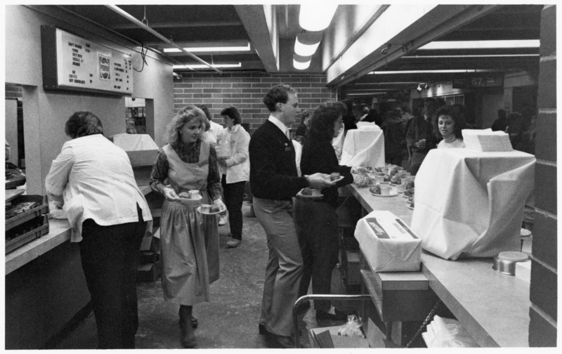 Black and white photograph of people carrying plates of food around a food concessions area.