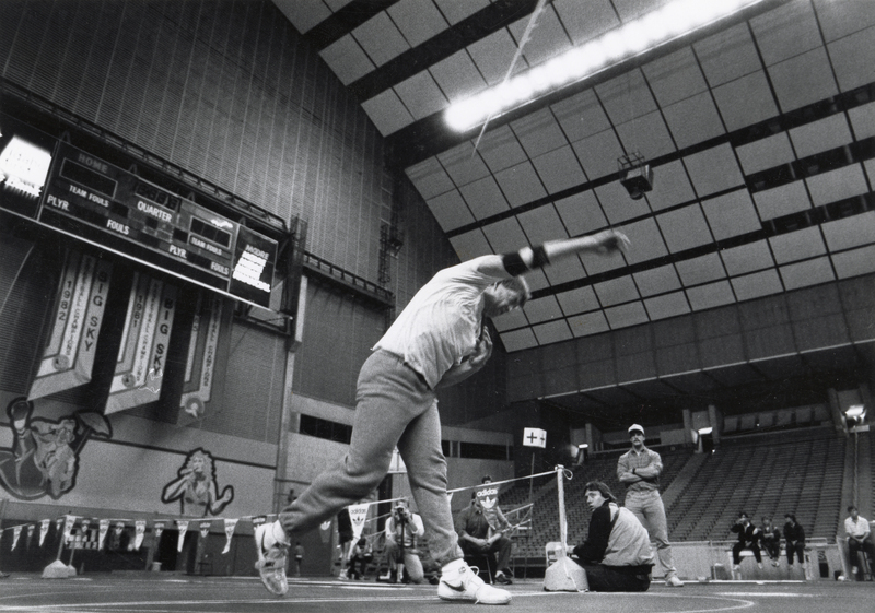 Black and white photograph of a javelin thrower standing on the field of an enclosed stadium. In the background are empty stands and several other people.