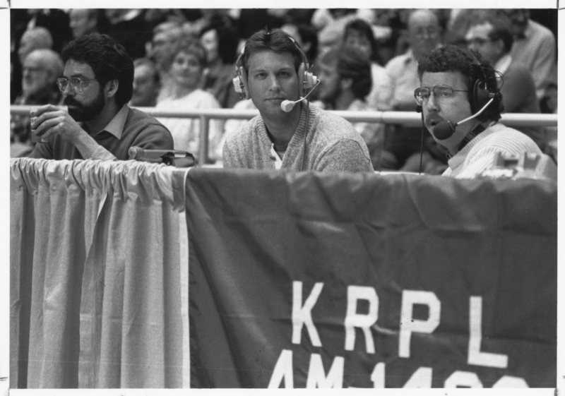 Black and white photograph of three men sitting at a table behind a low curtain, which reads "KRPL". In the background are spectators sitting in stands.