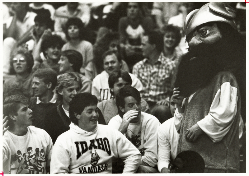 Black and white photograph of spectators sitting in stands interacting with a person in a mascot costume.