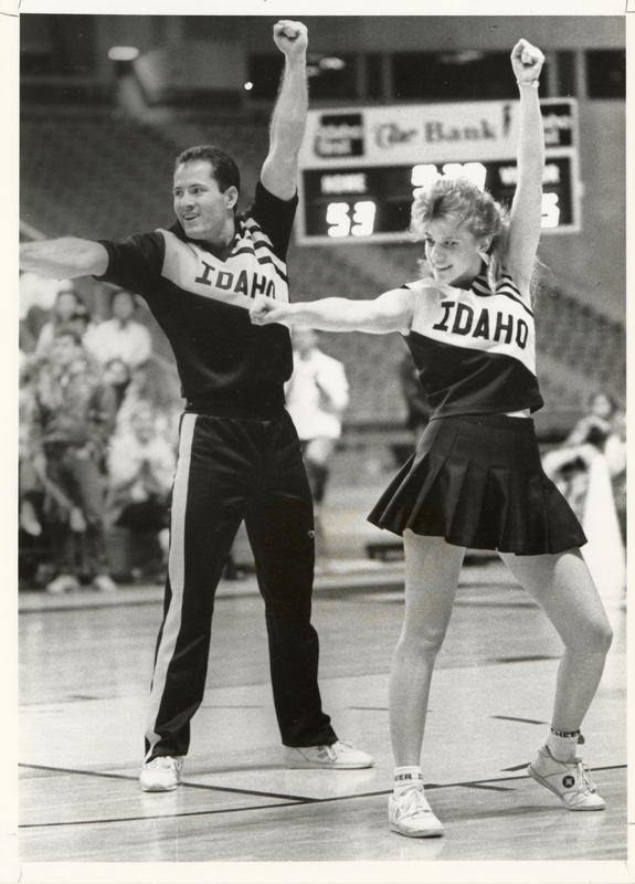 Black and white photograph of two people in cheerleader uniforms that read "IDAHO". In the background are spectators sitting in stands and a scoreboard.