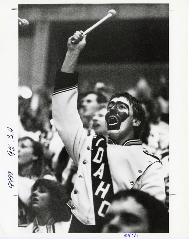 Black and white photograph of a person in a marching band uniform that reads "IDAHO." They hold a mallet and are surrounded by spectators in stands.