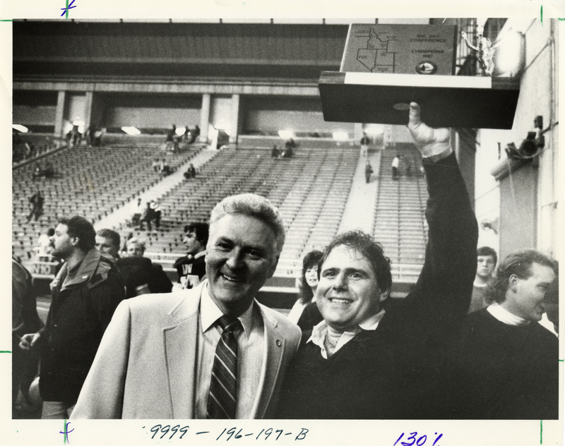 Black and white photograph of two men standing together, one holding a plaque that reads "BIG SKY CONFERENCE CHAMPIONS 1987". In the background are mostly empty stands.