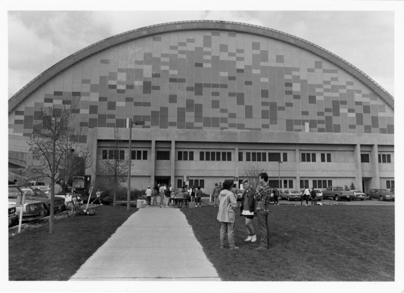 Black and white photograph of a building with an arched roof. In the foreground stand several people talking to each other.