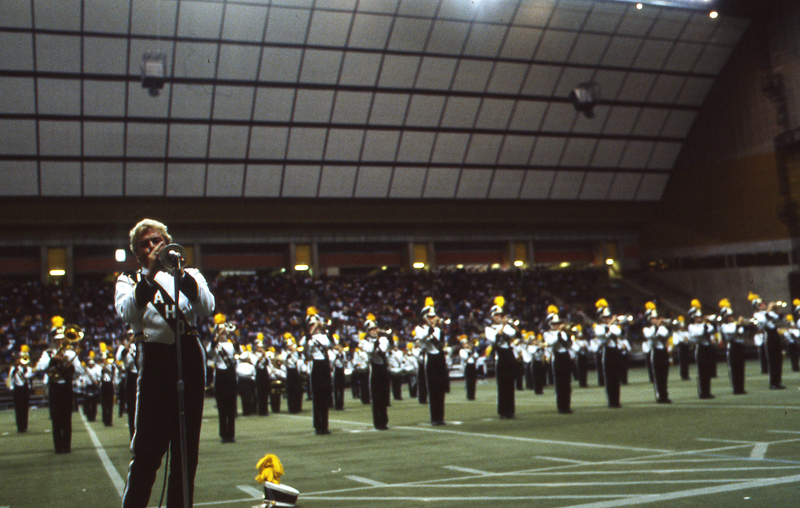 Color photograph of a trumpeter in a marching band uniform playing into a microphone. In the background are more marching band members and stands filled with people.