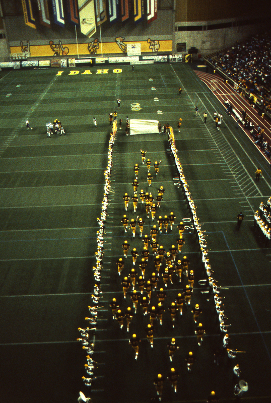 Color photograph of football players in black and yellow uniforms running onto a football field, flanked by members of a marching band.
