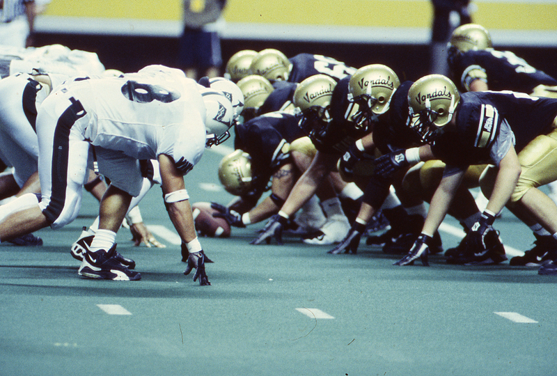 Color photograph of members of two football teams crouching and facing each other on a football field. One team wears white and black, the other wears gold and black.