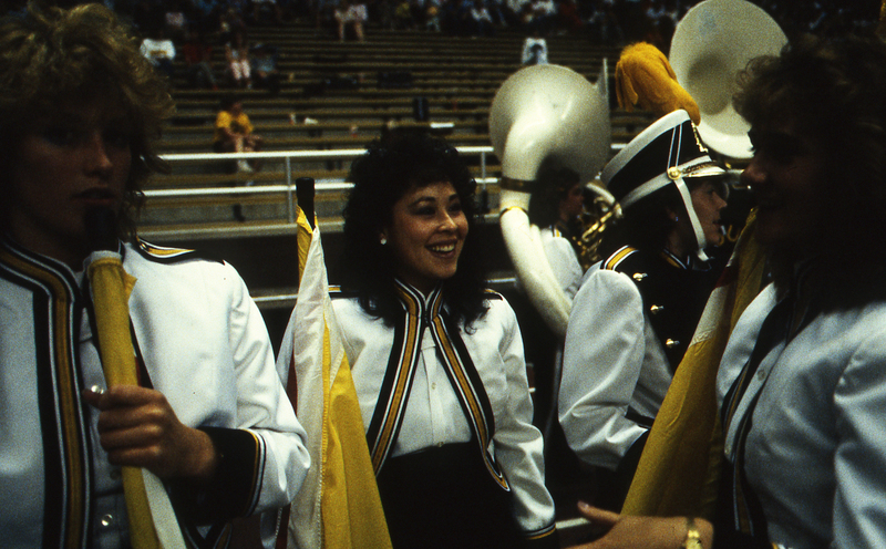 Color photograph of women wearing white, yellow, and black uniforms and holding white and yellow flags. In the background, people sit in stands.