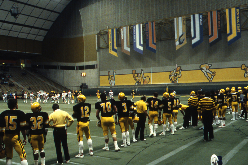 Color photograph of football players in black and yellow uniforms standing on the sidelines of an enclosed football field. In the background, football players face off against each other on the field.
