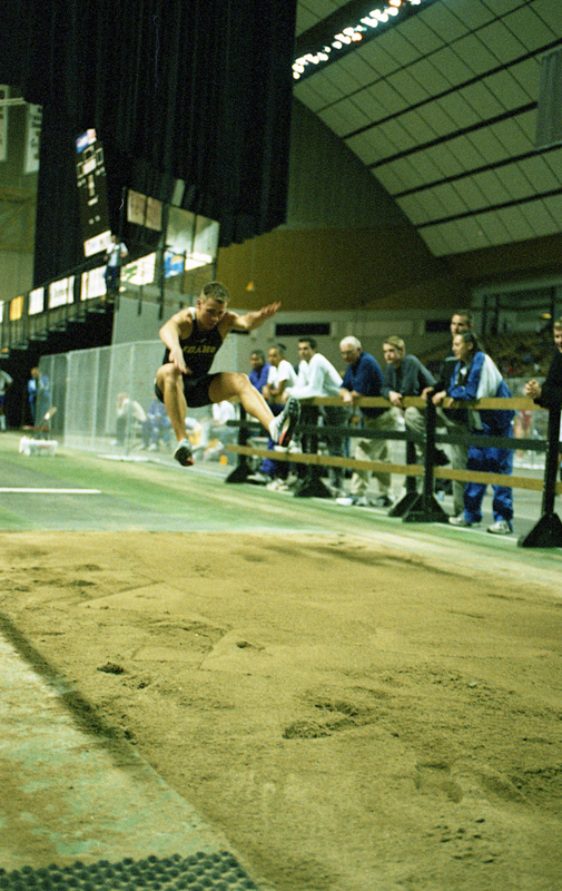 Color photograph of a man jumping into a sand pit while spectators watch from behind a wooden fence.