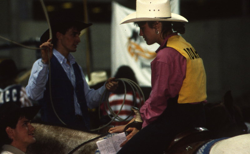 Color photograph of a woman riding a horse and interacting with a man standing nearby. The back of her vest reads "IDAHO".