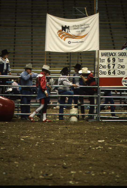 Color photograph of people standing along a metal fence on a dirt field inside of a stadium. A banner above them reads "Where Tradition Meets the Future University of Idaho".