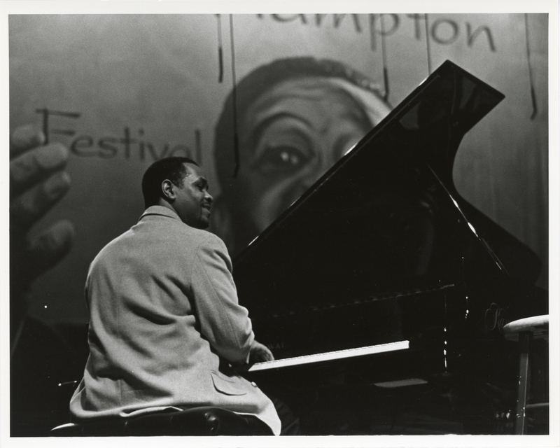 Black and white photograph of a man sitting at a piano. In the background is a large banner with a drawing of a man, partial text reading "Hampton Festival"