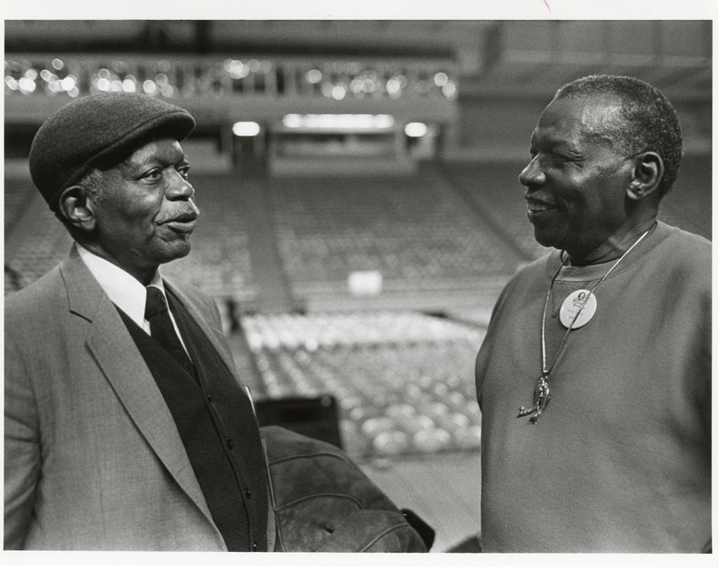 Black and white photograph of two men talking to each other. In the background are empty rows of chairs and stands.