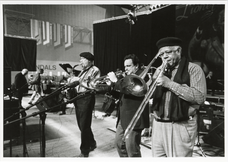 Black and white photograph of musicians playing saxophone, trumpet, and trombone on a stage.