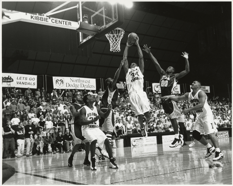 Black and white photograph of a men's basketball game in progress. Their uniforms read "IDAHO VANDALS" and "IONA". In the background, people sit or stand on stands.