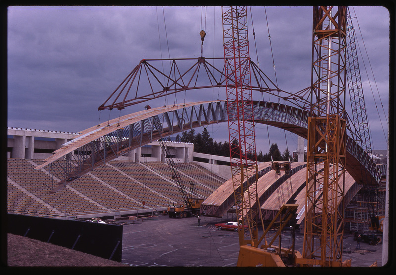 Color photograph of large wooden arches sitting on a stadium field and being maneuvered by large orange and red construction cranes.