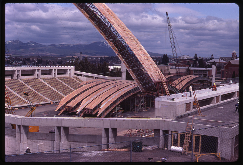 Color photograph of large wooden arches sitting on a stadium field and being maneuvered by large orange and red construction cranes.