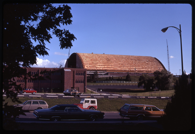 Color photograph of a domed building covered with plywood in a crosshatched pattern; in the foreground are a brick building and parked cars.