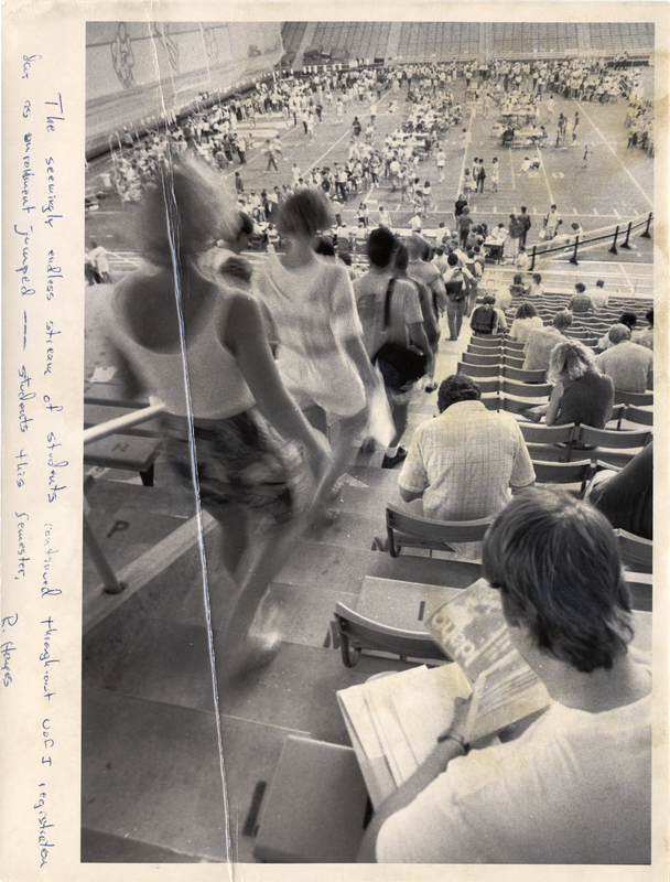 Black and white photograph of people walking down stadium stairs toward a football field where many other people mill about.
