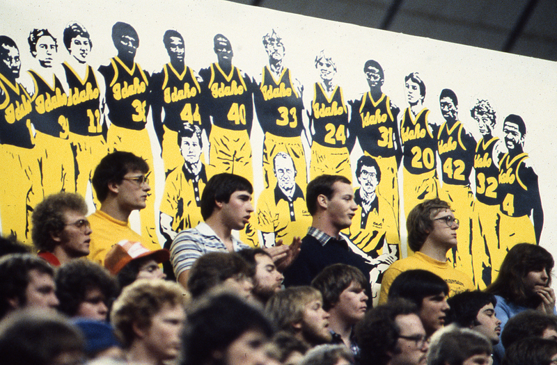 Color photograph of spectators standing or sitting in stands. Behind them is a black, white, and yellow drawing of basketball players and coaches lined up.