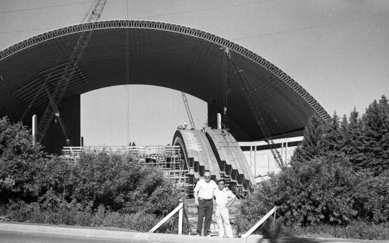 Two men stand near the construction site of a building with an arched roof, more wooden arches on the ground behind them.