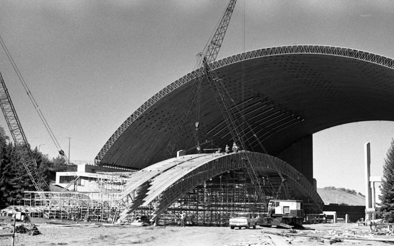Black and white photograph of an arched wooden roof under construction, surrounded by construction cranes. More wooden arches sit on the ground.
