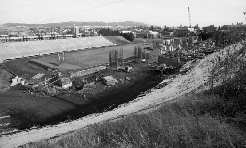 Black and white photograph of a horseshoe-shaped open air stadium with construction equipment on the field.